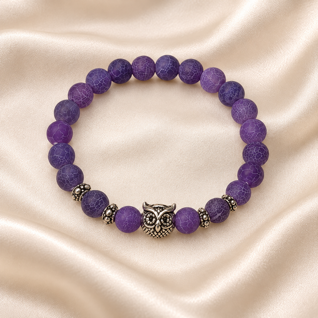 Purple Owl Bead Bracelet on pale champagne satin, showing matte purple crackle-pattern beads, a silver-tone owl centerpiece and decorative spacers.