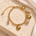 Seashell Treasures Charm Bracelet styled on ivory stone and linen, with three gold scallop charms and cream shell-look accents.