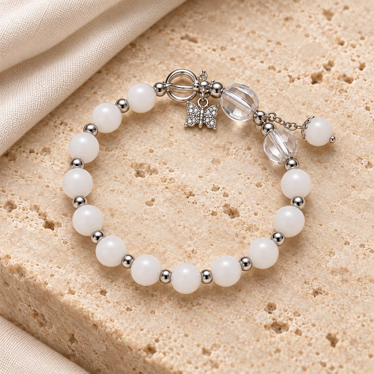 Snow White Bead Bracelet styled on ivory stone and linen, showing white beads, silver-tone spacers, clear accents and butterfly detail.