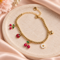Cherry Garden Charm Bracelet with gold-tone beads and cherry, ladybug, daisy, and heart charms displayed on a pale stone tray with blush satin.
