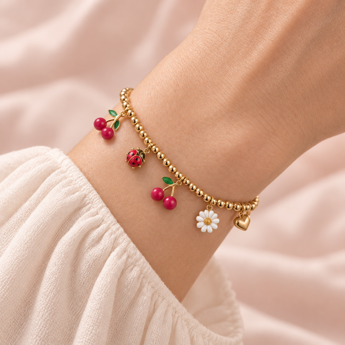 Close-up of the gold-tone Cherry Garden Charm Bracelet worn on a wrist, showing two cherry charms, a ladybug, white daisy, and heart.