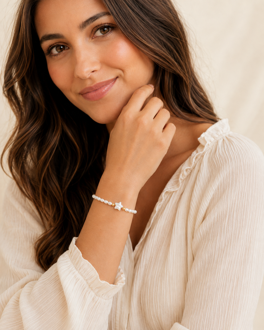 A woman with long brown hair wearing a delicate white beaded bracelet with a small gold star charm, posing in a cream-colored blouse against a soft beige background.