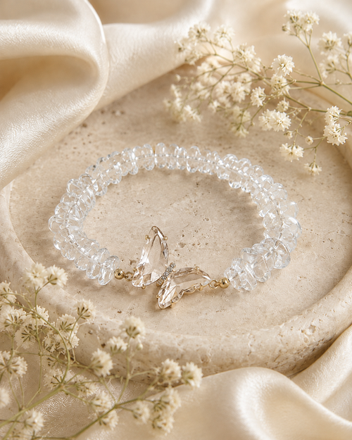 Clear crystal butterfly bracelet displayed on a neutral stone tray with soft satin fabric and floral styling.

