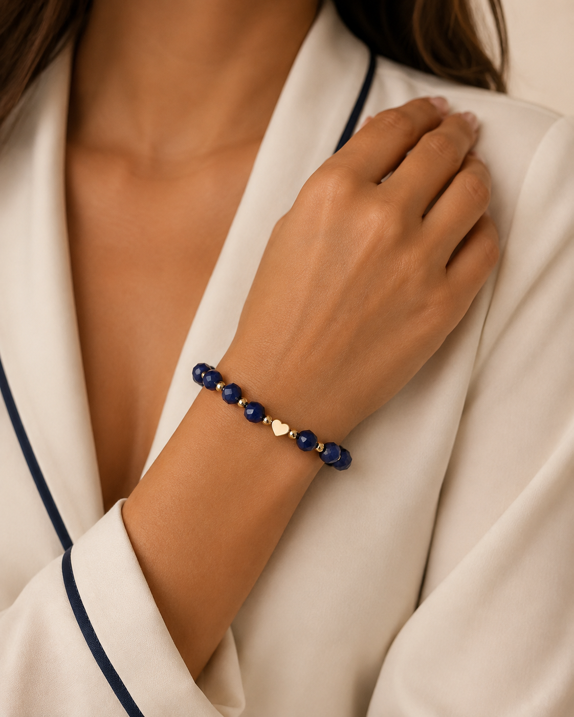 Model wearing a navy blue faceted bead bracelet with gold spacer beads and a small gold heart charm, styled with a cream and navy outfit.