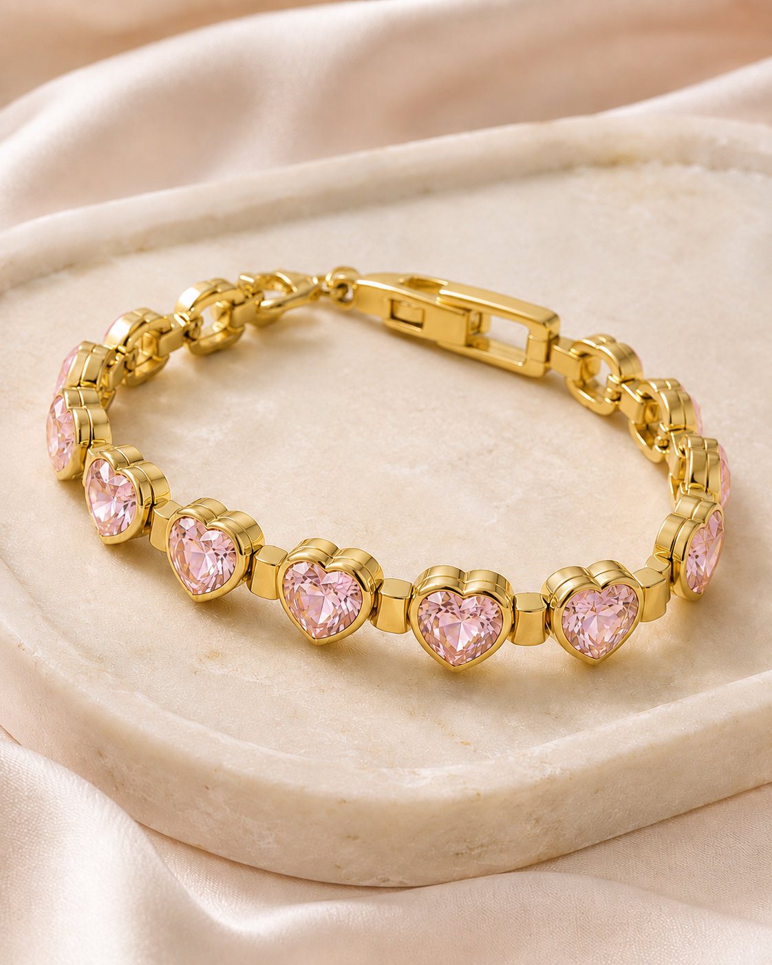 
Pink Heartstone Bracelet displayed on a neutral stone tray with satin fabric, gold-tone links, and sparkling pink heart details.
